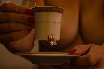 Primeiro plano da foto com uma xícara de café, e no plano posterior uma mulher nua, segurando a xícara, desfocada. A xícara e as mãos escondem os seios